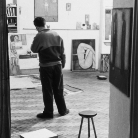 Mężczyzna stojący tyłem w przestrzeni pracowni. Obok niego stoi taboret. Pod ścianami i na podłodze znajdują się prace.