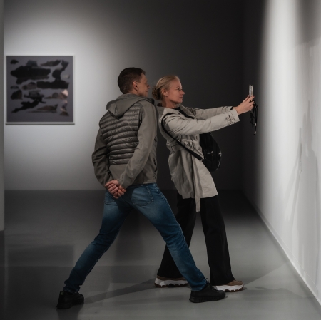 Kobieta i mężczyzna stoją blisko siebie w korytarzu galerii i wspólnie robią zdjęcie smartfonem oświetlonej ściennej ekspozycji. Mężczyzna w sportowej kamizelce i jeansach wychyla się mocno do przodu, a kobieta w jasnym płaszczu trzyma telefon w wyciągniętej dłoni.