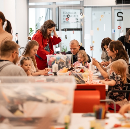 Gwarna sala warsztatowa wypełniona rodzinami z dziećmi, które siedzą przy stołach zastawionych plastikowymi pojemnikami z materiałami plastycznymi i klejem. W tle, za przeszklonymi drzwiami z napisem „EDUKACJA WARSZTATY”, wiszą na sznurkach kolorowe taśmy i kartki.