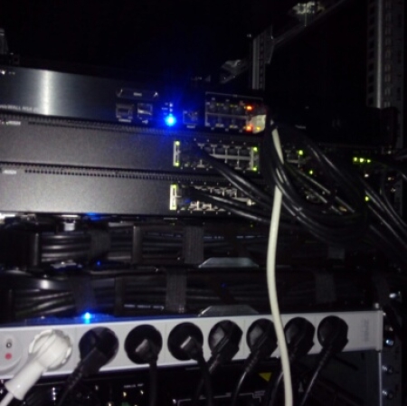 Zbliżenie na szafę serwerową w ciemnym pomieszczeniu, zawierającą kilka urządzeń sieciowych z podłączonymi kablami Ethernet i świecącymi diodami LED, a poniżej listwę zasilającą z wieloma wpiętymi przewodami.