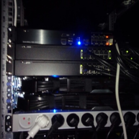Zbliżenie na szafę serwerową w ciemnym pomieszczeniu, zawierającą kilka urządzeń sieciowych z podłączonymi kablami Ethernet i świecącymi diodami LED, a poniżej listwę zasilającą z wieloma wpiętymi przewodami.