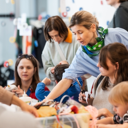 Półzbliżenie na kobietę w jasnoniebieskiej koszuli z zieloną apaszką w grochy, która pochyla się nad stołem i pomaga dziecku w pracy plastycznej. Wokół widać inne osoby pracujące przy stole oraz wiszącą nad nimi instalację z kolorowych taśm.
