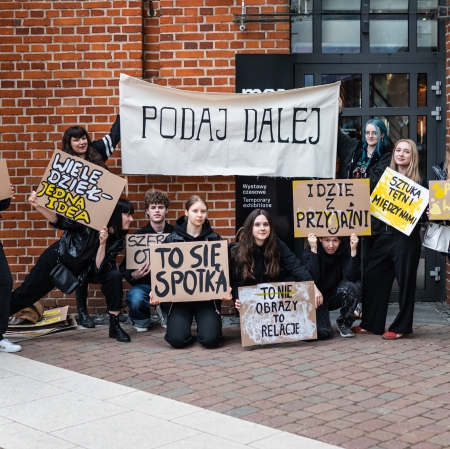 Grupa uśmiechniętej młodzieży pozuje na tle ściany z czerwonej cegły przed wejściem do budynku, trzymając ręcznie wypisane transparenty i kartony z hasłami takimi jak „PODAJ DALEJ” czy „TO NIE OBRAZY TO RELACJE”. Część osób stoi, a kilka kuca i klęczy na bruku, tworząc zgrany zespół.