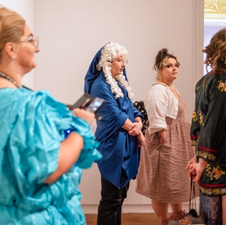 Grupa czterech osób w różnorodnych strojach i makijażach stoi w rzędzie w muzealnej sali, podczas gdy na pierwszym planie widać z profilu kobietę w jasnoniebieskiej, marszczonej kreacji.