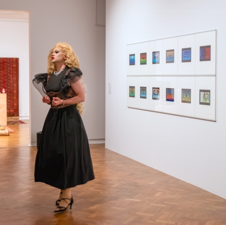 Drag queen w blond peruce i czarnej sukni przechodzi przez galerię sztuki, w której na ścianach wiszą oprawione w ramki kolorowe kwadraty, a w tle widać dywany i rzeźby.