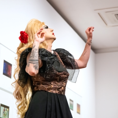 Widok z dołu na drag queen w blond peruce z czerwoną różą, która w pełnej ekspresji pozie wznosi ręce ku górze na tle białej ściany z galerią obrazów przedstawiających kasety magnetofonowe.