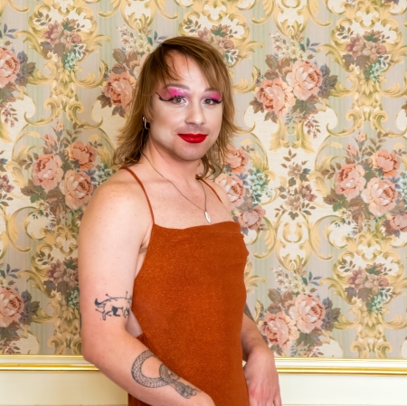 Portret osoby o brązowych włosach z wyrazistym makijażem, w tym jaskraworóżowym cieniem do powiek i czerwoną szminką, ubranej w pomarańczową sukienkę na ramiączkach, pozującej na tle tapety w kwiaty