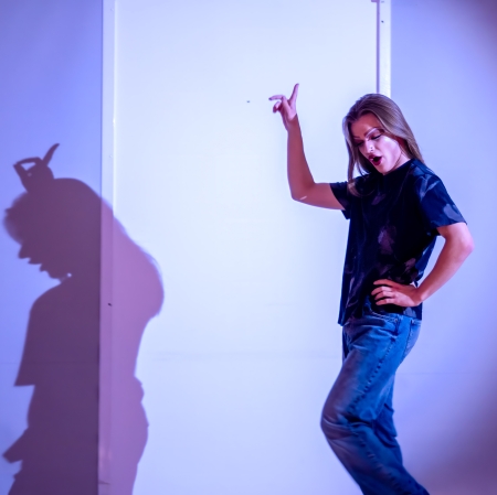 Osoba o długich, prostych włosach z mocnym makijażem tańczy z uniesioną ręką w niebieskim świetle reflektora, ubrana w ciemny t shirt i jeansy, podczas gdy na białej ścianie obok niej widoczny jest jej wyraźny cień.