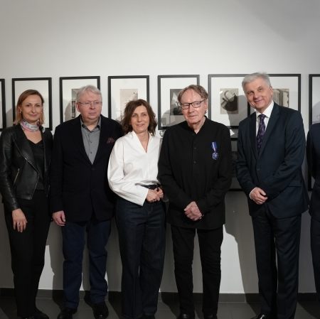 Grupa siedmiu elegancko ubranych osób pozuje do wspólnego zdjęcia na tle ściany z oprawionymi fotografiami.