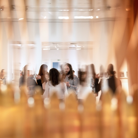 Artystyczne, rozmyte ujęcie wnętrza galerii sztuki, sfotografowane zza ażurowej lub przezroczystej przeszkody na pierwszym planie, która tworzy złote refleksy. W głębi sali, w świetle ostrości, widać rozmawiającą grupę ludzi, w tym mężczyznę z brodą i dredami.
