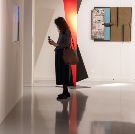 Kobieta stoi na gładkiej, błyszczącej posadzce galerii i przegląda coś w smartfonie na tle geometrycznych dzieł sztuki – dużej, czerwono-czarnej rzeźby oraz wiszącego na ścianie panelu.