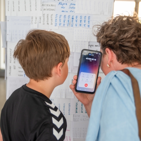 Kobieta i chłopiec stoją blisko tablicy z ogłoszeniami, sprawdzając numer telefonu wyświetlony na ekranie smartfona.