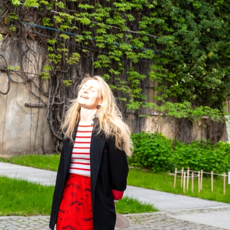 Uśmiechnięta blondynka w czerwonej spódnicy i czarnej marynarce stoi na brukowanym dziedzińcu na tle ściany gęsto porośniętej zielonym bluszczem.