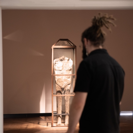 Mężczyzna z długimi dredami spiętymi w kok patrzy od tyłu na stojącą w głębi sali muzeum rzeźbę ludzkiej sylwetki, zamkniętą w szklanej gablocie.
