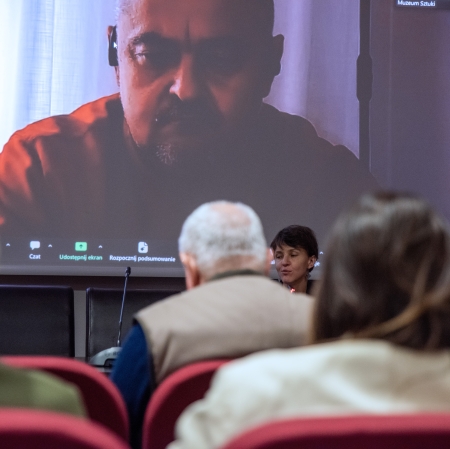 Zdjęcie zrobione z perspektywy widowni skupia się na twarzy kobiety z panelu dyskusyjnego oraz na monumentalnym wizerunku mężczyzny na ekranie, podczas gdy sylwetki osób w pierwszym planie pozostają rozmyte.