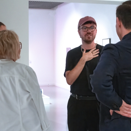 Młody mężczyzna w okularach i różowej czapce z daszkiem stoi w galerii sztuki, gestykulując dłonią przy klatce piersiowej podczas rozmowy z grupą osób stojących tyłem do obiektywu.