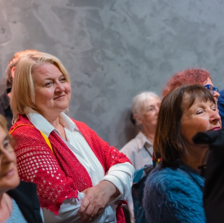 Bliskie ujęcie zasłuchanej publiczności na tle szarej ściany, pośród której wyróżnia się uśmiechnięta kobieta w białej bluzce i czerwonym, ażurowym swetrze.