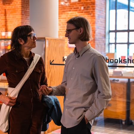 Kobieta w okularach i brązowym kombinezonie rozmawia z młodym mężczyzną w jasnej koszuli w przestrzeni muzealnej księgarni z napisem „bookshop ms²”.