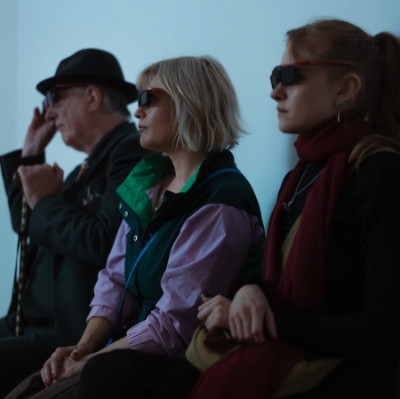 Trzy osoby siedzą profilem w rzędzie przy ścianie w chłodnym, niebieskawym świetle i oglądają prezentację, mając na sobie specjalne okulary (prawdopodobnie 3D).