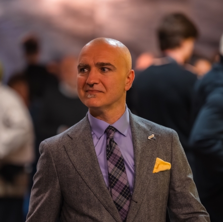 Portret eleganckiego, łysego mężczyzny w szarym, wełnianym garniturze, fioletowej koszuli i wzorzystym krawacie z żółtą poszetką w kieszeni. Mężczyzna patrzy w bok z lekkim, zagadkowym uśmiechem na tle rozmytego tłumu gości.