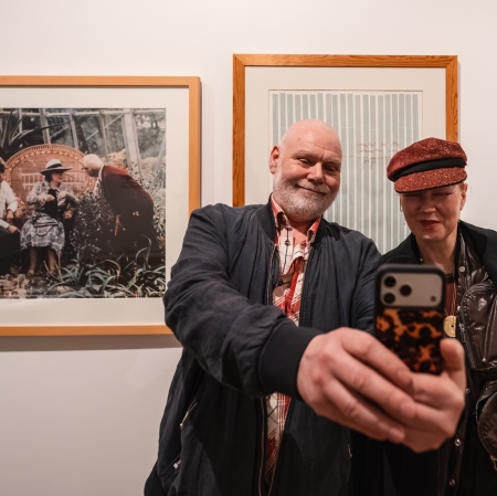 Mężczyzna z brodą i kobieta w błyszczącym, bordowym kaszkiecie robią sobie wspólne zdjęcie („selfie”) smartfonem w galerii sztuki. W tle na białej ścianie wiszą dwa duże dzieła sztuki w jasnych, drewnianych ramach – fotografia oraz grafika.