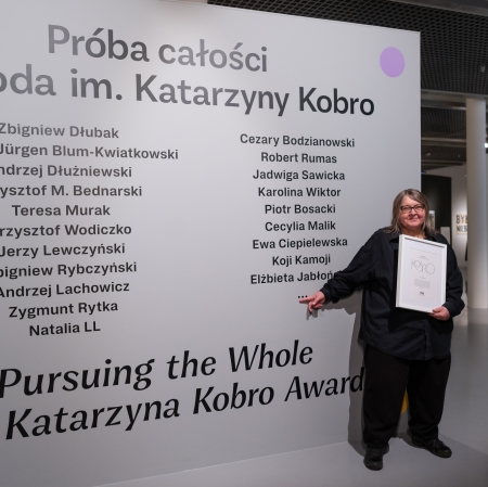 Osoba stojąca w przestrzeni wystawy, wskazuje dłonią na element znajdujący się na ścianie