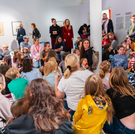 Duża grupa dzieci wraz z opiekunami siedzi blisko siebie na podłodze galerii, uczestnicząc w prelekcji prowadzonej przez stojących wokół edukatorów.