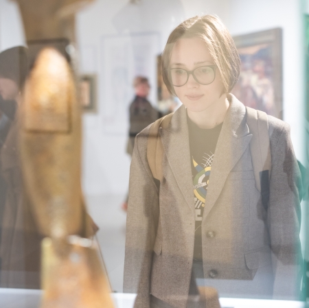Młoda osoba w okularach i szarej marynarce patrzy z zaciekawieniem na rzeźbę wyeksponowaną w gablotcie. Scena rozgrywa się w jasnym wnętrzu galerii sztuki lub muzeum, a w rozmytym tle widać inne dzieła oraz zwiedzających.