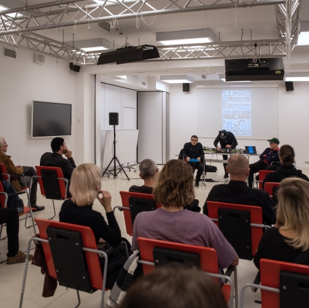 In the foreground, a group of people sit on red chairs. All are facing the men sitting in the center of the room.