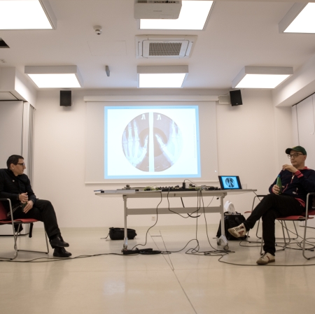 On either side of the image, we see two people seated, with a table with DJ equipment between them. In the background, a screen with graphics is displayed.