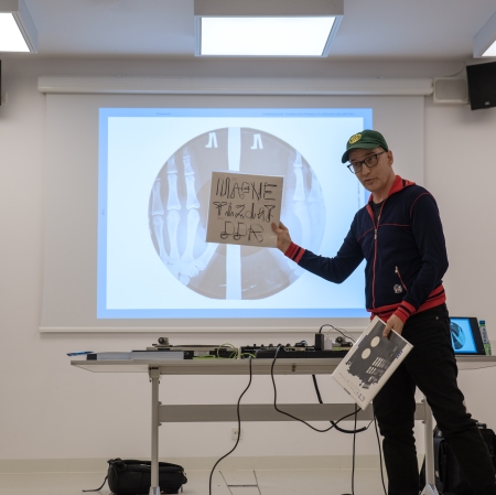 A man wearing a cap and glasses holds a vinyl record in his right hand. In the background is a screen with graphics displayed.