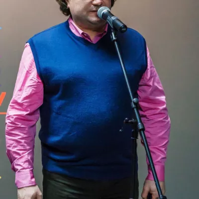 Na zdjęciu pan w kamizelce, berecie i z teczką mówi do mikrofonu.