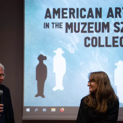 Dwie osoby stoją na tle wyświetlanej nieieskiej grafiki z napisem "American artists in the Muzeum Sztuki Collecton"