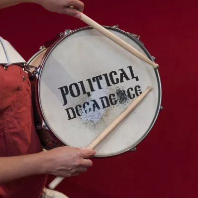 Fotografia przedstawia biały werbel z napisem ”Political decadence”, zdjęcie wykonane jest z góry. Osoba ubrana w czerwony kombinezon trzyma werbel i pałki perkusyjne, tło również jest czerwone.  