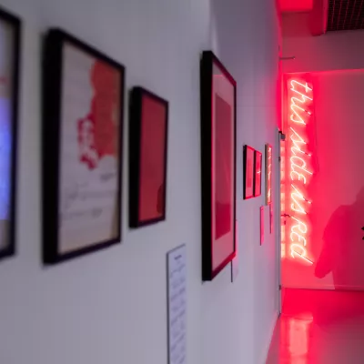 Przestrzeń wystawy, na ścianie po lewej stronie wiszą na ścianie grafiki, na ścianie na wprost zawieszony jest czerwony neon, obok niego tyłem do aparatu stoi człowiek ubrany w czarny płaszcz
