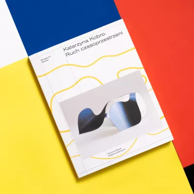 Na tle białych, niebieskich, żółtych, czerwonych i czarnych prostokątów ukośnie leży książka Katarzyna Kobro. Ruch czasoprzestrzeni, na okładce rzeźba o nieregularnhych kształtach