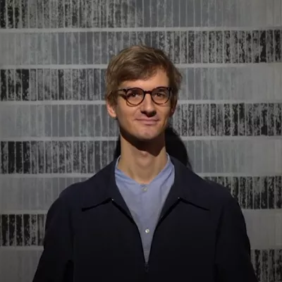 Na pierwszym planie znajduje się kurator, Jakub Gawkowski, w tle widoczna czarno-biała praca będąca częścią wystawy