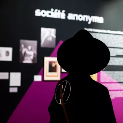 Na zdjęciu kobieta w czarnym kapeluszu na głowie, odwrócona tyłem do fotografa spogląda na napis "Société anonyme"