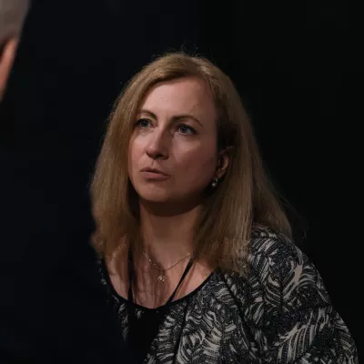 Kobieta o blond włosach do ramion stoi na tle czarnej ściany i rozmawia z osobą stojącą przed nią.