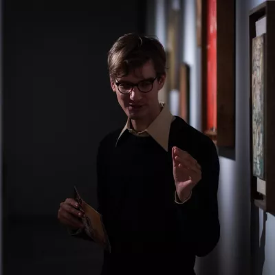 Kurator Jakub Gawkowski stoi obok wiszących na ścianie obrazów Rosenstein i Williamsa. W jednej z dłoni trzyma broszurę wystawy, drugą gestykuluje, opowiadając coś publiczności.