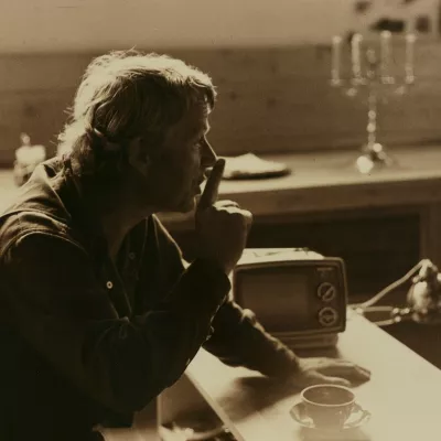 Na zdjęciu znajduje się Roman Opałka. Siedzi on przy biurku i przykłada palec do ust. Na fotografię nałożona jest sepia. 