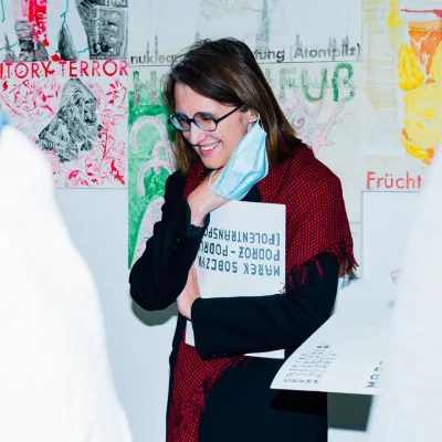 Na zdjęciu kobieta patrzy w dół, uśmiecha się, prawą ręką podtrzymuje maseczkę w dłoni trzyma katalog do wystawy.