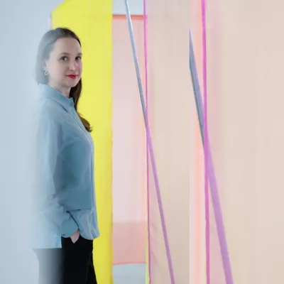 Młoda kobieta ubrana w jasno niebieską koszulę, obok fragmenty instalacji w postaci delikatnej tkaniny w kolorach różowym i żółtym. Kobieta patrzy na widza i uśmiecha się. 