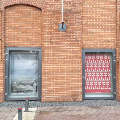 The graphic is a photo of two windows. On the left, there is the artwork by Anna Bąk, on the right, there is a project by Witek Grethengier. The photo of Anna Bąk's installation shows a battered sofa in a gray, metallic box. Witek Grethengier's artwork consists of red folk cutouts.