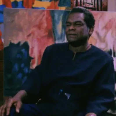 Aubrey Williams sitting in the studio surrounded by his paintings. He is wearing a navy pullover 