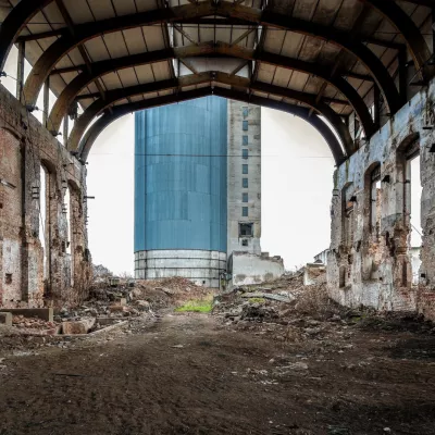 The photo shows a ruined, industrial, red-brick building. In the background, there is a modern skyscraper.