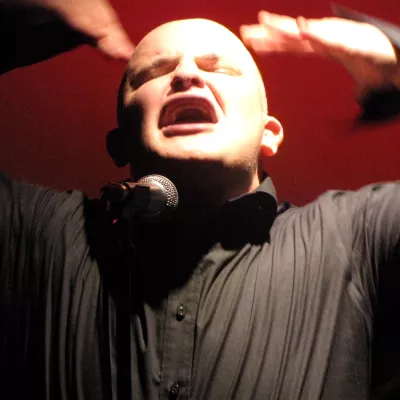 mężczyzna ma przed sobą mikrofon, unosi ręce do góry, ma otwarte usta