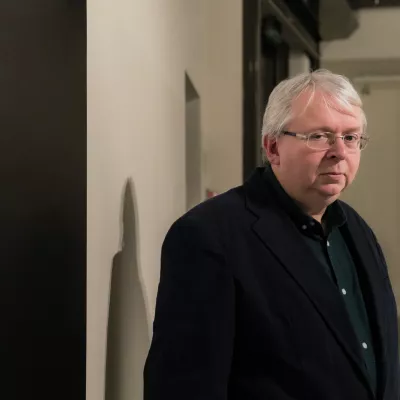 Portret mężczyzny w okularach, w ciemnym garniturze. Mężczyzna patrzy w stronę obiektywu, po lewo fragment ciemnej ściany.