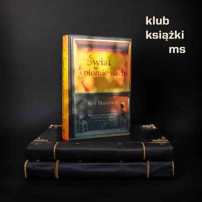 Książka Siri Hustvedt pt. „Świat w płomieniach” stojąca na dwóch czarnych, skórzanych tomach w ozdobnej oprawie. Całość przedstawiona na czarnym tle z jasnym napisem „klub książki ms” w prawym górnym rogu.