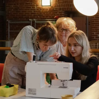Młoda kobieta w czarnej bluzce obsługuje maszynę do szycia marki Janome pod okiem starszej kobiety w okularach i innej młodej kobiety w paski. Wszystkie trzy pochylają się nad maszyną w oświetlonym pomieszczeniu o industrialnym wystroju.